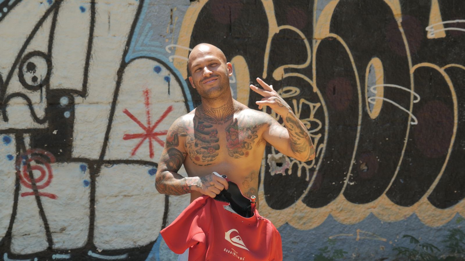 Marcelo grins in front of a graffiti wall, holding his red impact vest