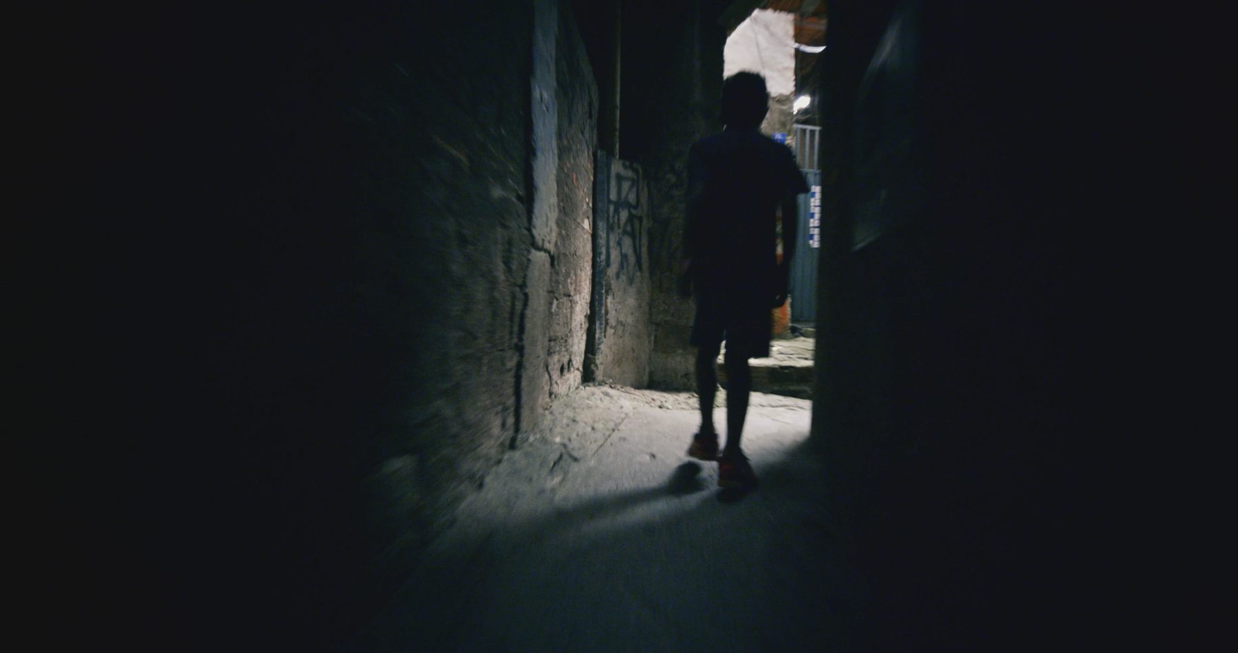 A boy in silhouette walks into a dark alley towards a pool of light