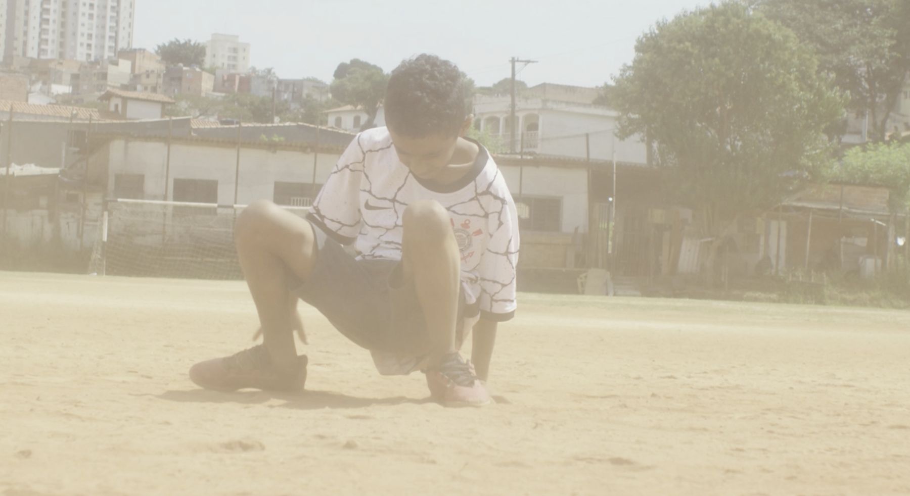 A boy crouches on a hazy, sunlit dirt pitch