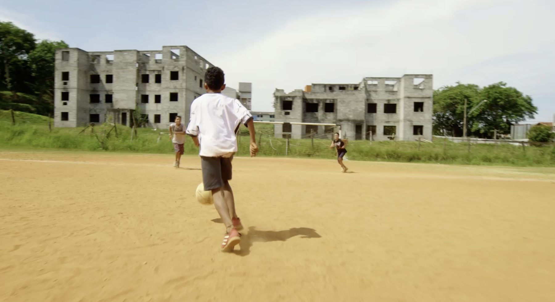 A boy dribbles a football across a dirt pitch past unfinished buildings