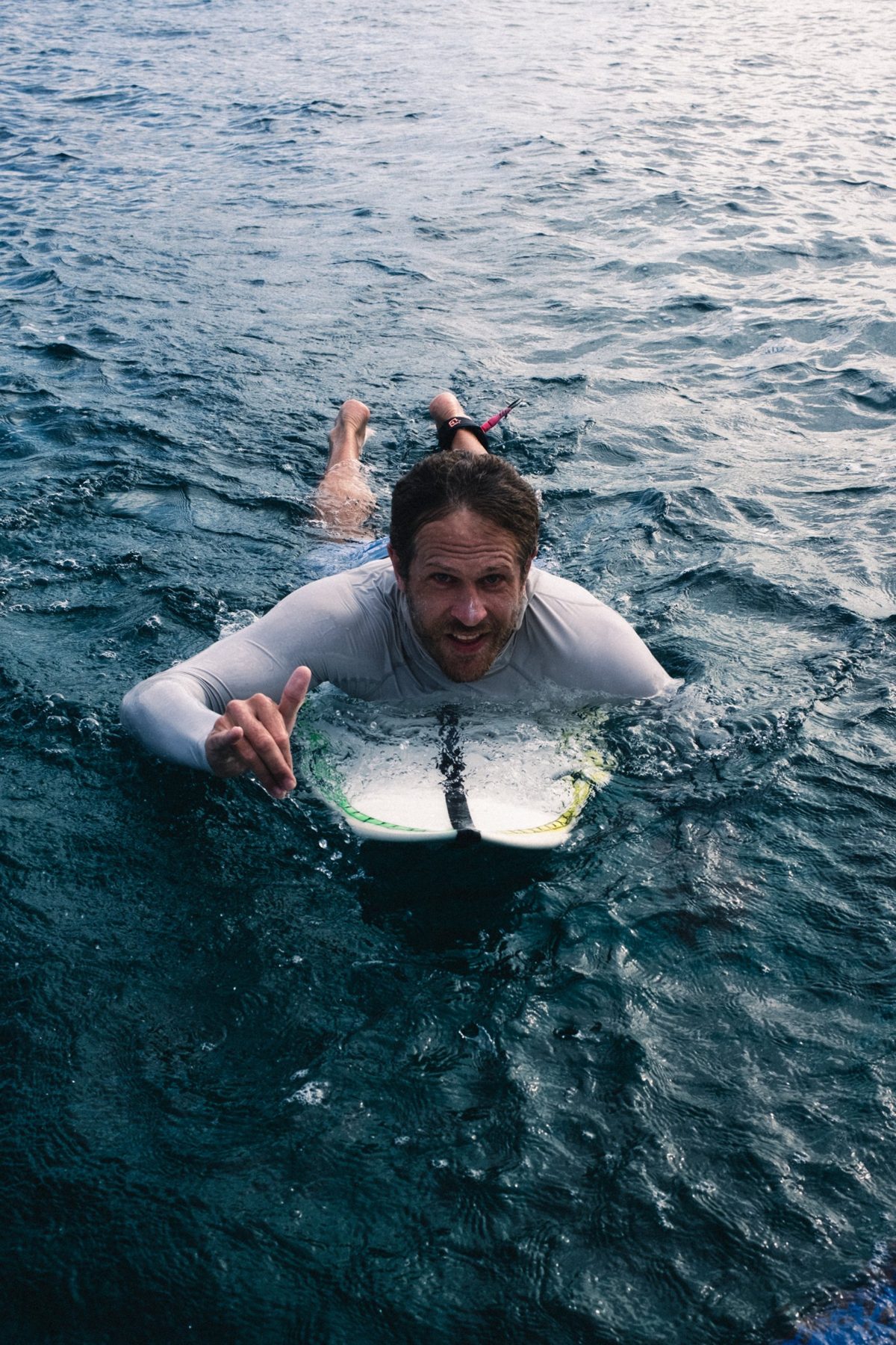 Nick paddles towards camera, grinning and throwing a shaka