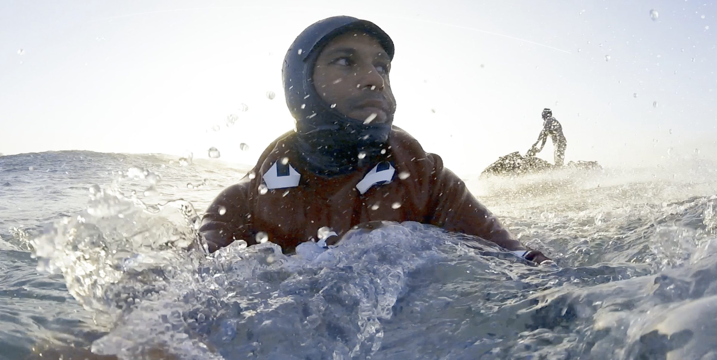 Marcelo in the water at Nazaré, hood up, a jet ski waiting behind him