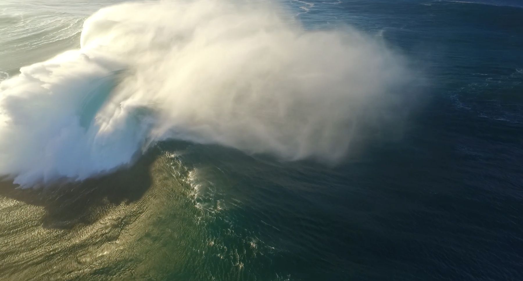 From above: spray tears off the lip of a giant wave in low sun
