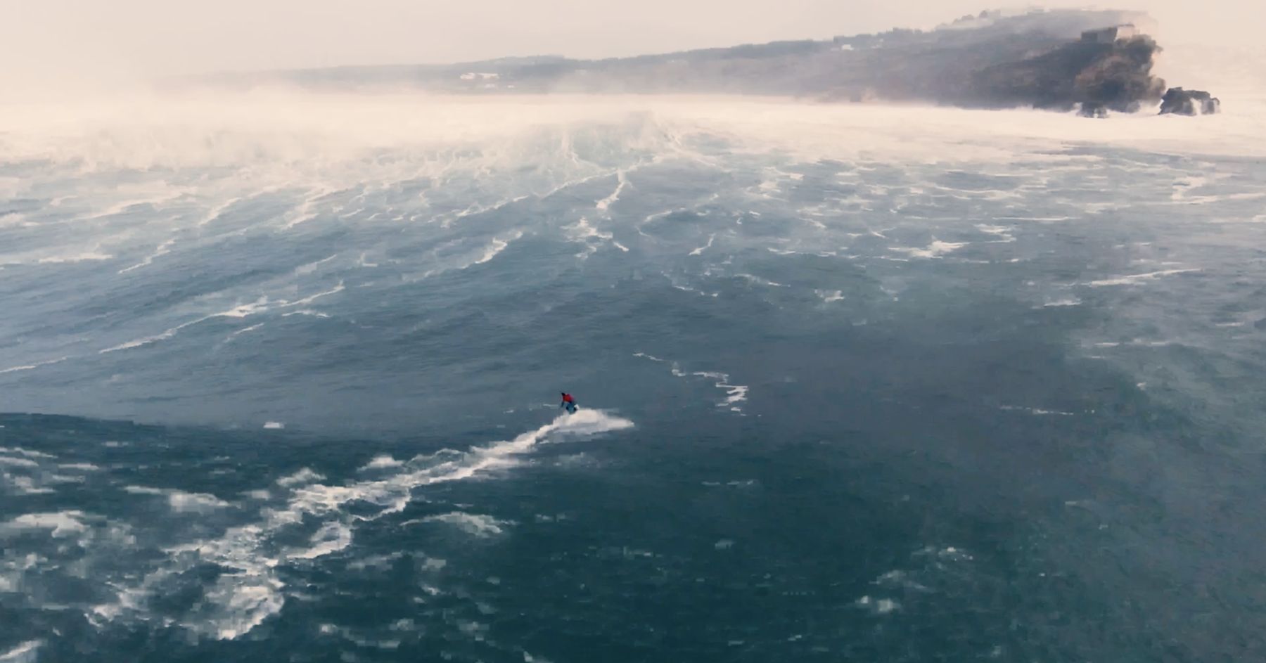 A lone surfer in red rides through wind-whipped water below the cliffs of Nazaré